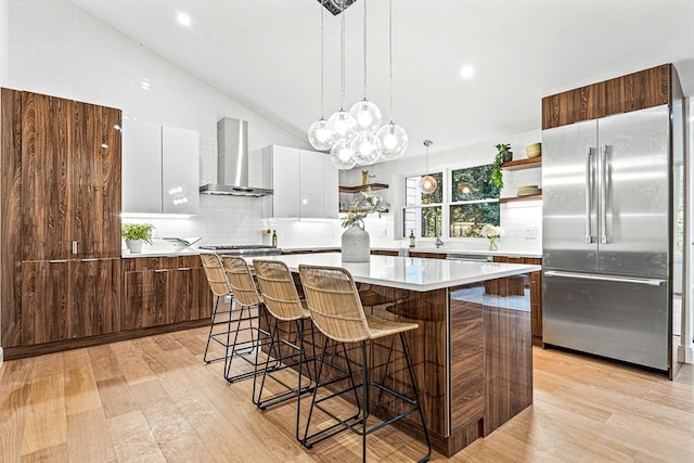 kitchen with a kitchen island, wall chimney range hood, white cabinetry, built in fridge, and vaulted ceiling