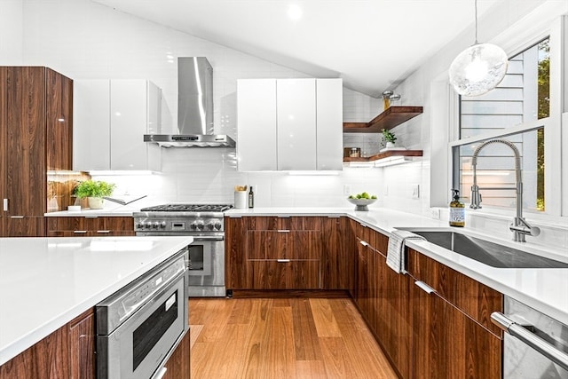 kitchen featuring hanging light fixtures, vaulted ceiling, white cabinets, stainless steel appliances, and wall chimney range hood