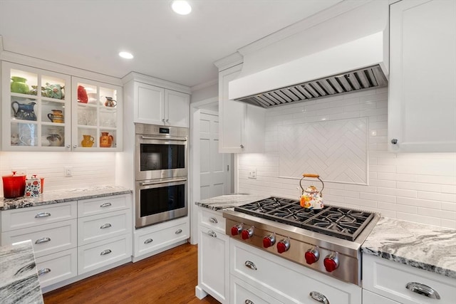 kitchen with dark wood finished floors, appliances with stainless steel finishes, glass insert cabinets, white cabinets, and wall chimney range hood