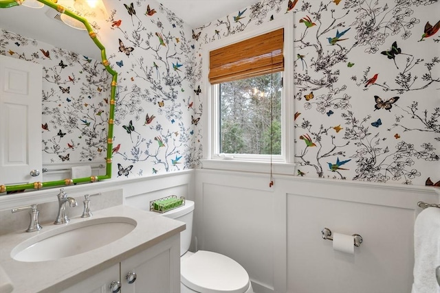 half bathroom featuring toilet, wallpapered walls, vanity, and wainscoting