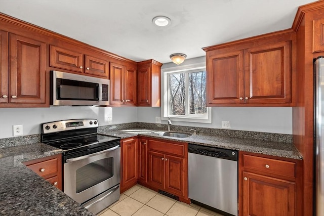 kitchen with brown cabinets, a sink, dark stone countertops, appliances with stainless steel finishes, and light tile patterned flooring