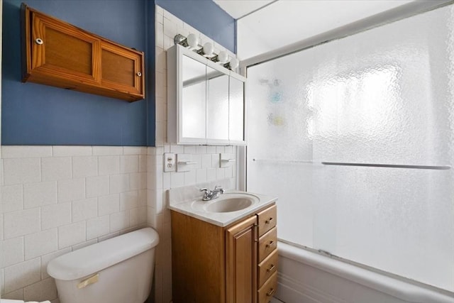 full bathroom featuring tile walls, bath / shower combo with glass door, toilet, vanity, and wainscoting