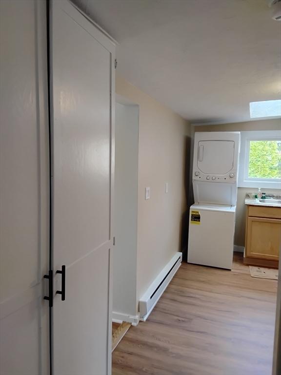 clothes washing area featuring light wood-type flooring, a baseboard radiator, stacked washer and clothes dryer, and a skylight