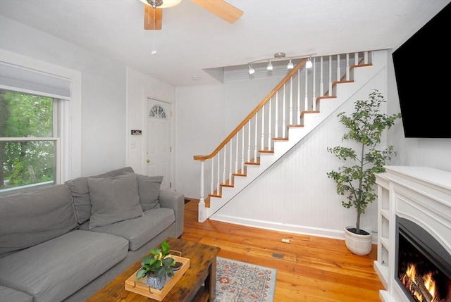 living room featuring stairs, hardwood / wood-style flooring, baseboards, a lit fireplace, and a ceiling fan