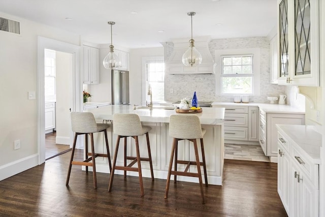 kitchen featuring dark wood-type flooring, stainless steel fridge, an island with sink, decorative light fixtures, and white cabinets