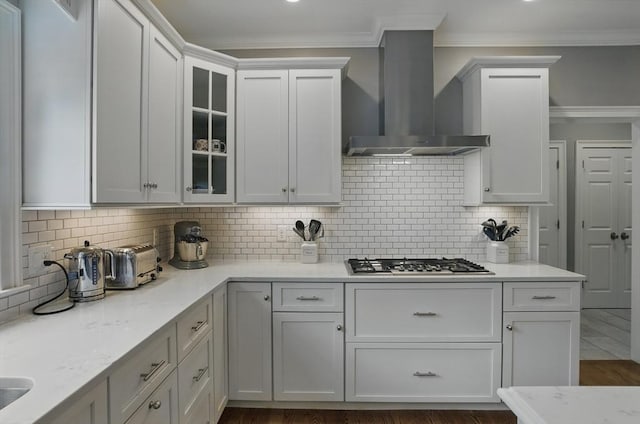 kitchen with crown molding, glass insert cabinets, wall chimney exhaust hood, light stone counters, and stainless steel gas stovetop