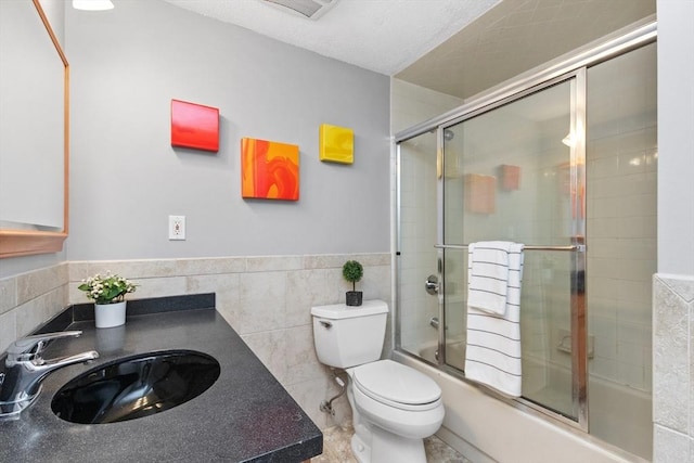 full bath with vanity, toilet, shower / bath combination with glass door, tile walls, and a wainscoted wall