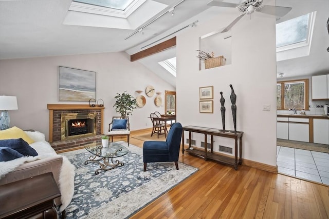 living room featuring vaulted ceiling with skylight, ceiling fan, rail lighting, light wood-type flooring, and a brick fireplace