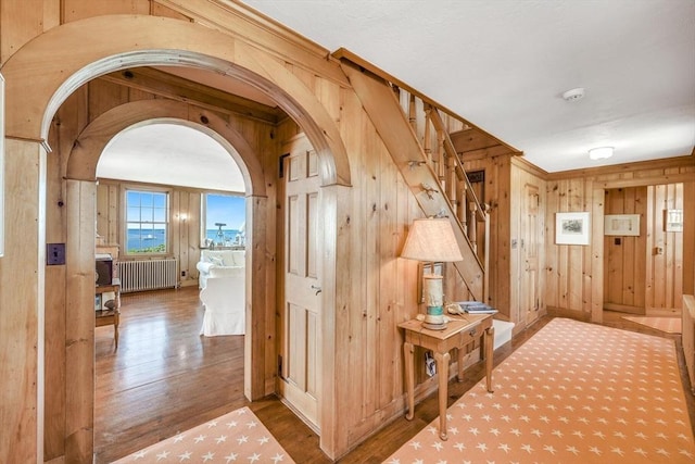 hallway with arched walkways, radiator, wood finished floors, stairs, and wood walls