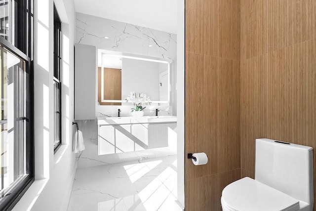 bathroom with marble tiled floors, toilet, double vanity, and stone wall