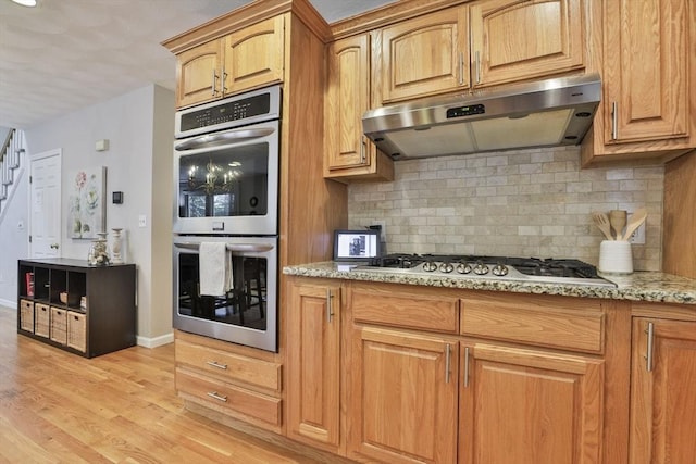 kitchen featuring decorative backsplash, appliances with stainless steel finishes, light stone counters, light wood-style floors, and under cabinet range hood