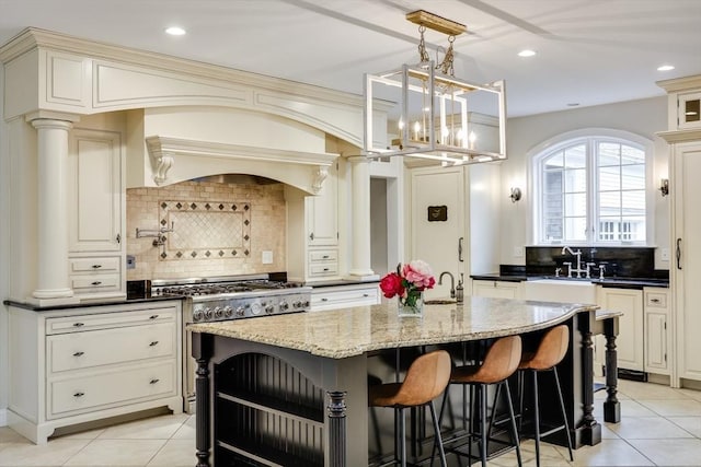 kitchen featuring backsplash, decorative columns, a kitchen island with sink, and dark stone counters