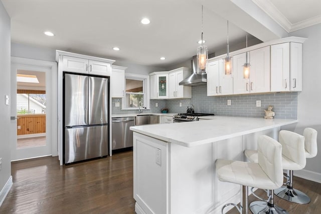 kitchen with light countertops, wall chimney exhaust hood, stainless steel appliances, a peninsula, and ornamental molding