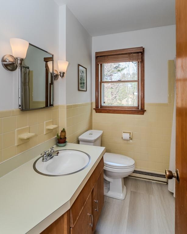 bathroom featuring wood finished floors, toilet, vanity, a baseboard heating unit, and tile walls
