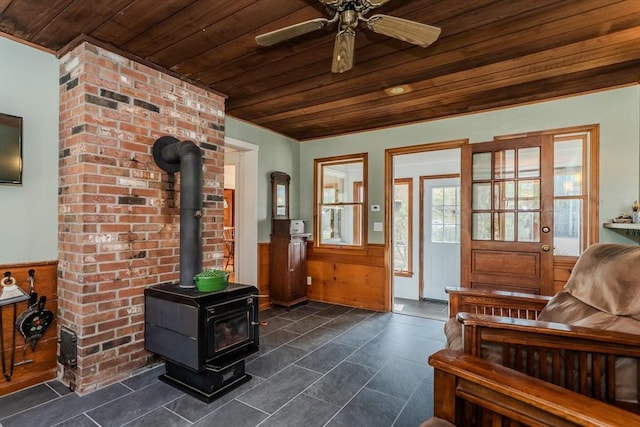 living area with a wood stove, wooden ceiling, a ceiling fan, and a wainscoted wall