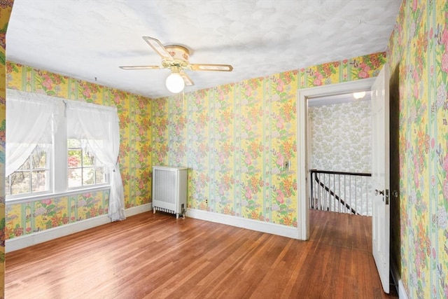 empty room featuring wood finished floors, baseboards, ceiling fan, radiator, and wallpapered walls