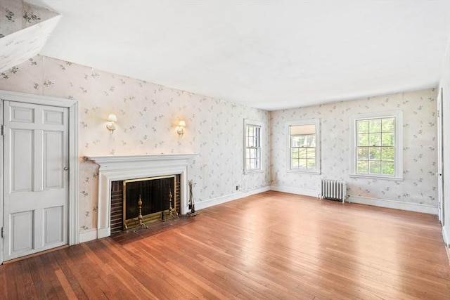 unfurnished living room with radiator heating unit, wood-type flooring, wallpapered walls, a fireplace with flush hearth, and baseboards