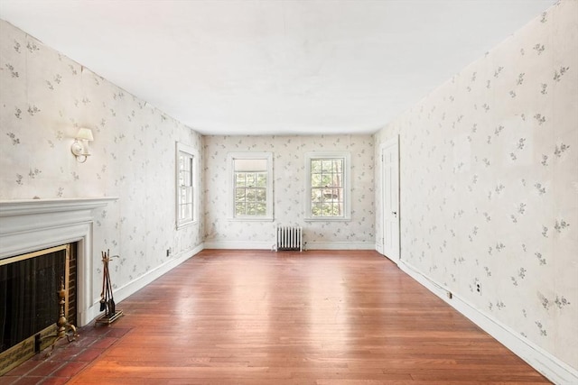 unfurnished living room with radiator, wood-type flooring, wallpapered walls, a fireplace with flush hearth, and baseboards