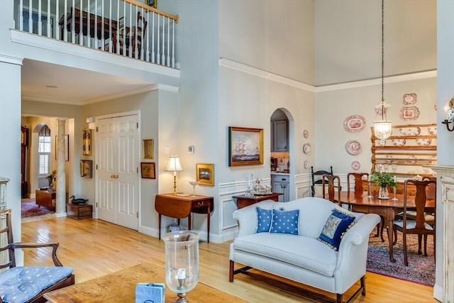 living area featuring arched walkways, a towering ceiling, light wood-style floors, ornamental molding, and decorative columns