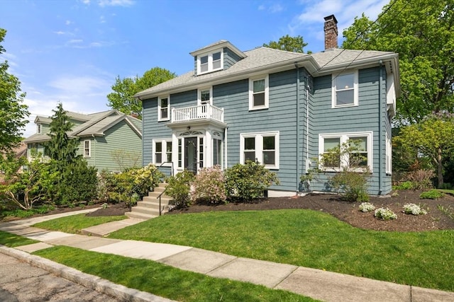 view of front of home with a balcony, a front yard, roof with shingles, and a chimney