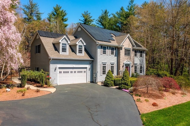 colonial house with an attached garage, driveway, solar panels, and a shingled roof