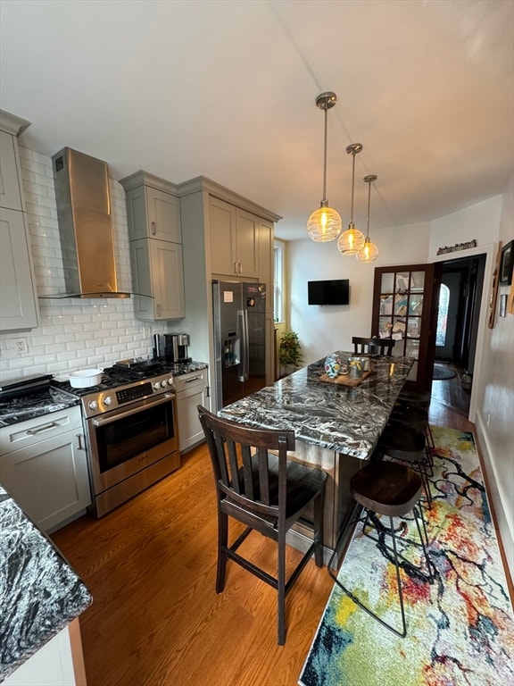 kitchen with wood-type flooring, dark stone counters, appliances with stainless steel finishes, hanging light fixtures, and wall chimney exhaust hood