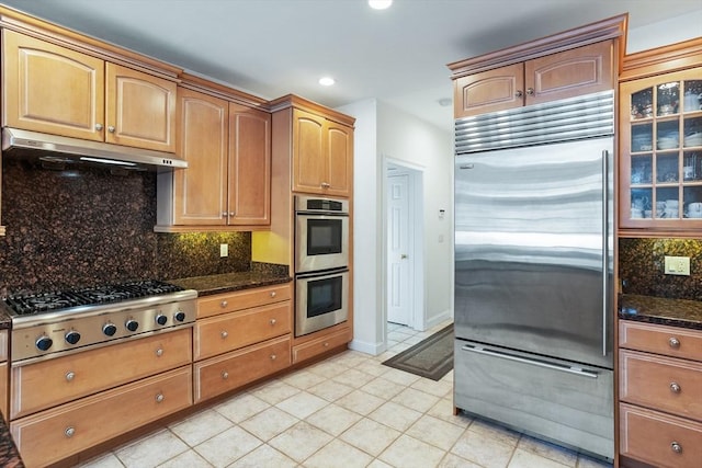 kitchen with under cabinet range hood, appliances with stainless steel finishes, backsplash, and dark stone countertops