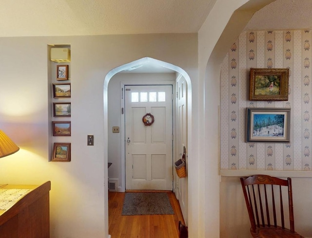 foyer featuring a textured ceiling, arched walkways, wood finished floors, visible vents, and wallpapered walls