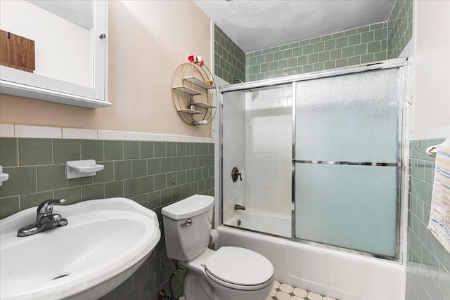 full bath with a sink, tile walls, combined bath / shower with glass door, toilet, and a wainscoted wall