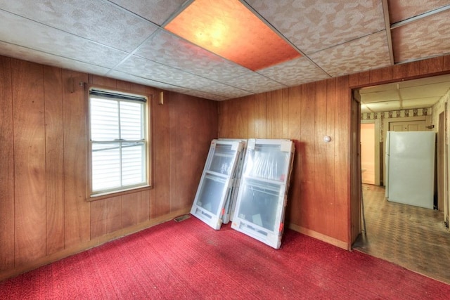 empty room with carpet floors, wooden walls, baseboards, and a paneled ceiling