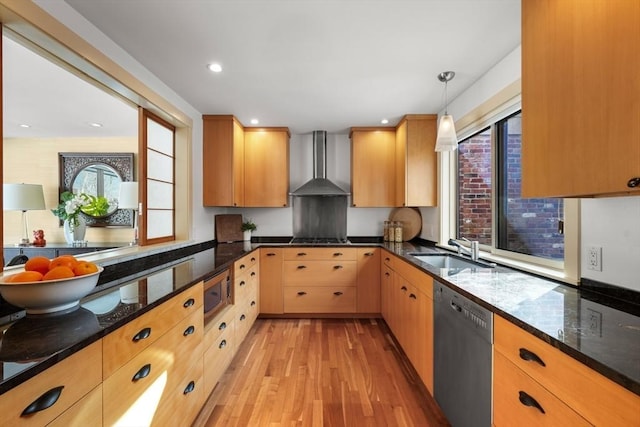 kitchen with light wood finished floors, dark stone countertops, stainless steel appliances, wall chimney range hood, and a sink