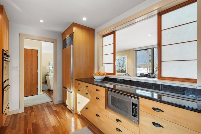 kitchen featuring dark stone countertops, built in appliances, light brown cabinetry, light wood-type flooring, and recessed lighting