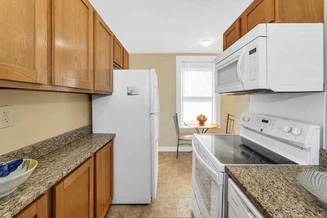 kitchen featuring baseboards, white appliances, brown cabinetry, dark stone counters, and light tile patterned flooring