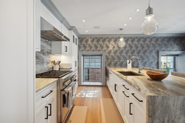 kitchen with sink, wall chimney exhaust hood, stainless steel stove, pendant lighting, and white cabinets