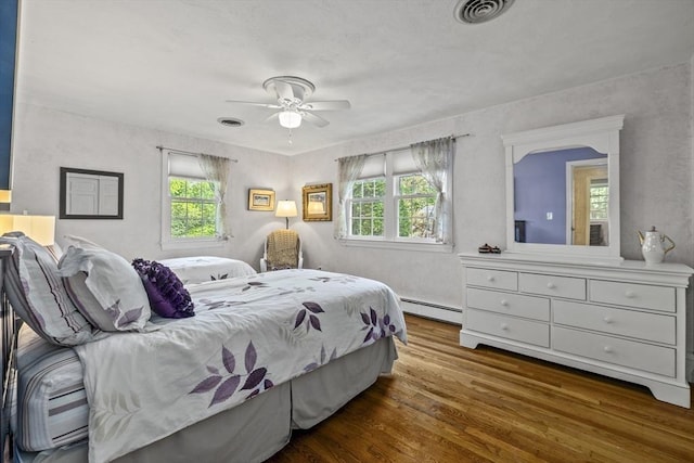 bedroom featuring a baseboard heating unit, multiple windows, wood finished floors, and ceiling fan