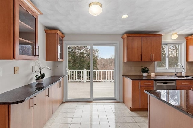 kitchen with light tile patterned floors, a sink, stainless steel dishwasher, dark stone countertops, and glass insert cabinets