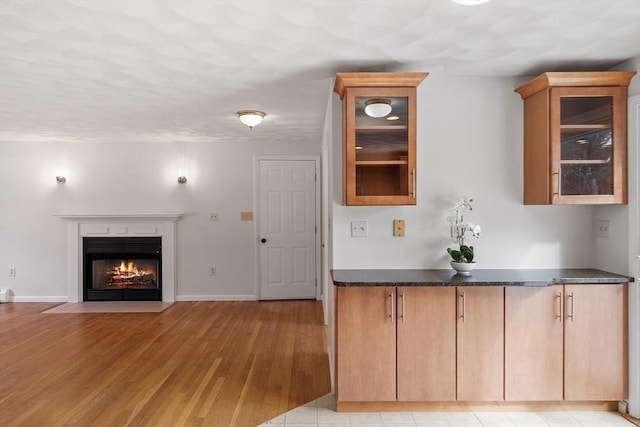 kitchen featuring light wood-style flooring, a fireplace with flush hearth, baseboards, dark countertops, and glass insert cabinets