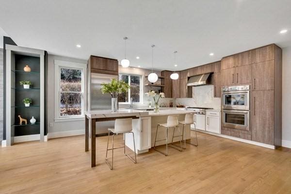 kitchen featuring wall chimney range hood, light wood-type flooring, a kitchen breakfast bar, and appliances with stainless steel finishes