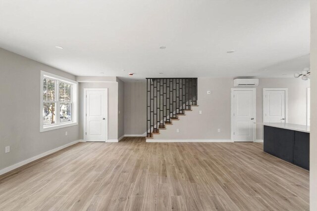 unfurnished living room with a wall mounted air conditioner, baseboards, light wood finished floors, and stairway