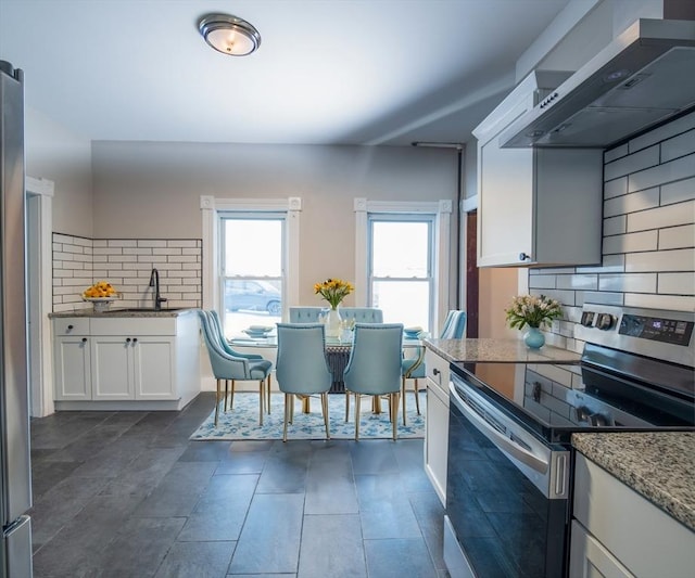 kitchen featuring wall chimney exhaust hood, backsplash, stainless steel electric stove, white cabinetry, and a sink