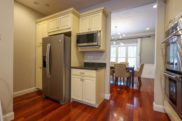 kitchen with a notable chandelier, stainless steel appliances, dark wood-type flooring, cream cabinetry, and dark stone counters