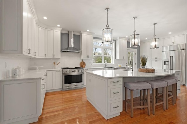 kitchen with premium appliances, wall chimney exhaust hood, a kitchen breakfast bar, a kitchen island with sink, and light countertops