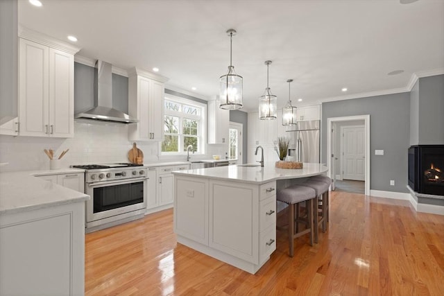 kitchen with light wood finished floors, appliances with stainless steel finishes, ornamental molding, white cabinetry, and wall chimney range hood