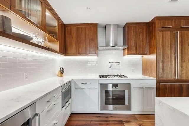 kitchen with dark wood-style floors, decorative backsplash, glass insert cabinets, built in appliances, and wall chimney range hood