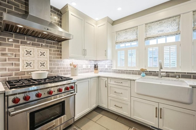 kitchen with wall chimney exhaust hood, luxury range, a sink, light tile patterned floors, and backsplash