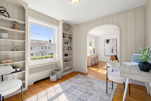 home office featuring radiator heating unit, arched walkways, built in shelves, and wood finished floors