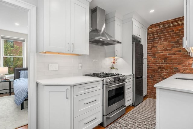 kitchen with appliances with stainless steel finishes, wall chimney range hood, light countertops, white cabinets, and brick wall