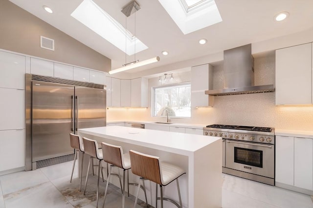 kitchen featuring high end appliances, wall chimney exhaust hood, lofted ceiling with skylight, and white cabinetry