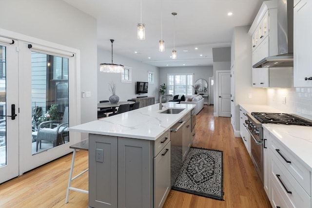 kitchen featuring wall chimney exhaust hood, sink, decorative light fixtures, light stone countertops, and a kitchen island with sink