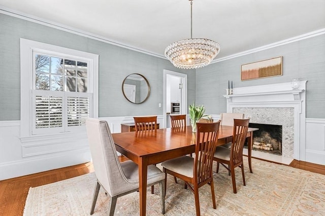 dining space featuring a tiled fireplace, ornamental molding, wainscoting, a chandelier, and light wood-type flooring